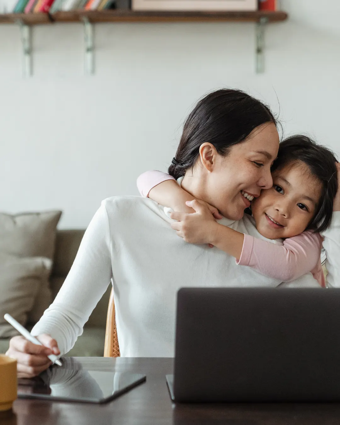 A mother working at a laptop while her daughter hugs her